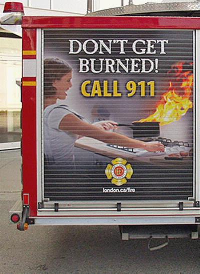 A billboard on a London fire truck that depicts a woman moving a pot, that has caught on fire, off the stove reads ‘Don’t get burned! Call 9-1-1.’ 