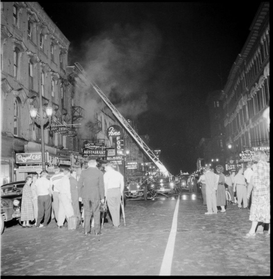 People gathering the street to watch the ladder truck extended up in to the building building