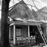 Fire fighters working from a ladder and on the front porch of a home with light smoke showing