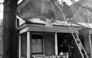 Fire fighters working from a ladder and on the front porch of a home with light smoke showing