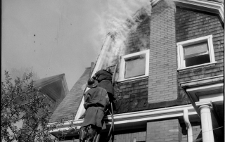 Two Fire Fighters working on a ladder to extinguish a fire within the wall and attic of the second floor of a home.