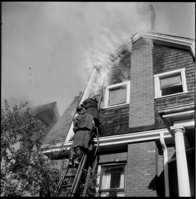 Two Fire Fighters working on a ladder to extinguish a fire within the wall and attic of the second floor of a home.