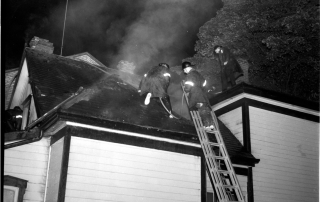 Three fire fighters on a roof extinguishing fire in the roof and attic