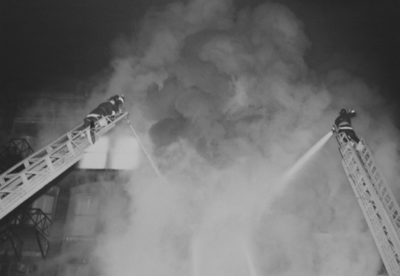 Two large aerial ladders set-up with as firefighter on each spraying water on the very large fire through the smoke.