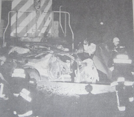 A nighttime scene shows firefighters and emergency personnel standing beside a heavily damaged car that was struck by a freight train. The front of the CPR locomotive fills the background with its striped pattern illuminated by scene lighting. First responders examine the crushed vehicle, with equipment and reflective gear visible in the dark surroundings.