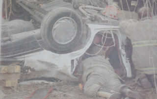 An overturned car rests on its roof at the bottom of an embankment as firefighters lie on the ground working beneath it to free trapped occupants. The vehicle is stabilized with cribbing, and rescue personnel in full gear are using hand tools to access the interior. Debris and branches surround the scene, indicating the car slid off a gravel road and struck a tree.