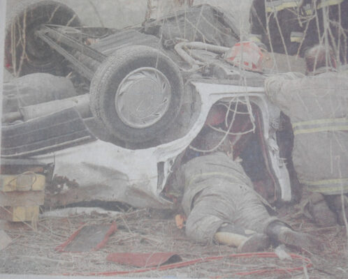An overturned car rests on its roof at the bottom of an embankment as firefighters lie on the ground working beneath it to free trapped occupants. The vehicle is stabilized with cribbing, and rescue personnel in full gear are using hand tools to access the interior. Debris and branches surround the scene, indicating the car slid off a gravel road and struck a tree.