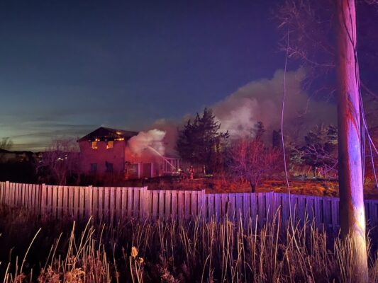 Far away photo of fire fighters spraying water in a home that has flames and smoke coming from the second storey roof line.