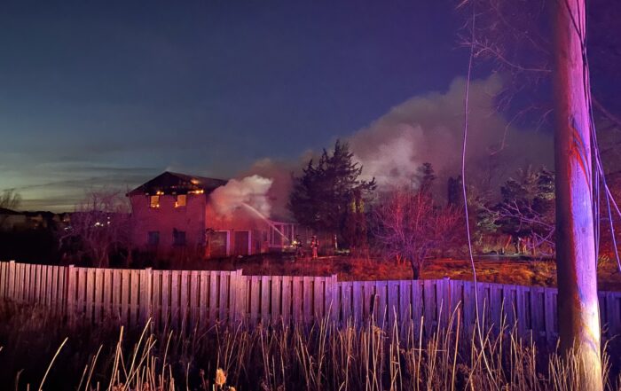 Far away photo of fire fighters spraying water in a home that has flames and smoke coming from the second storey roof line.