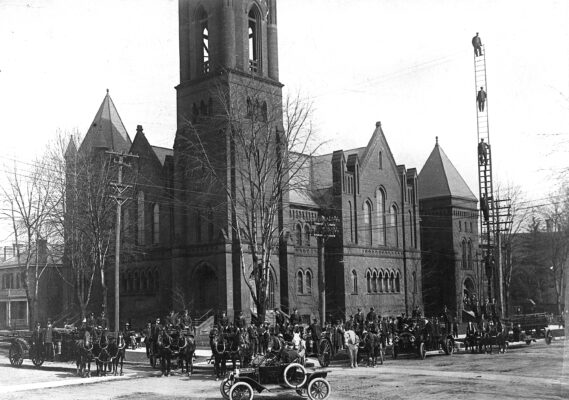Historic 1914 photograph showing a lineup of horse-drawn fire wagons, early motorized fire engines, and uniformed firefighters posed in front of a large stone church with tall towers and arched windows. A tall wooden extension ladder stands at the right side of the image with several firefighters positioned on its rungs. Bare trees and dirt roads indicate an early 20th-century streetscape.