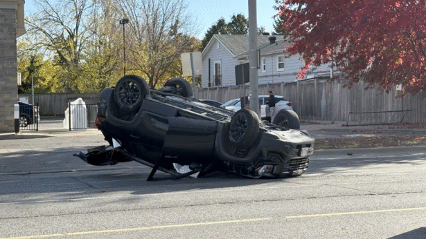 A black SUV lies flipped upside down in the middle of a city street, its roof crushed and undercarriage exposed. Debris is scattered around the vehicle, and a utility pole stands nearby. In the background, houses, a wooden fence, and autumn trees with red and yellow leaves are visible, along with a few bystanders observing the scene.