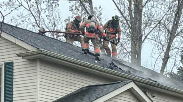 Three firefighters wearing full protective gear and air packs stand on the roof of a house, using tools to open a burned section where light smoke is escaping. The home’s shingles are damaged near the peak, and bare trees rise behind the scene, indicating an active ventilation operation during a structure fire.