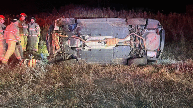 Firefighters in turnout gear stand next to a vehicle lying on its side in a grassy ditch at night, with the underside of the car exposed and emergency lights illuminating the scene.