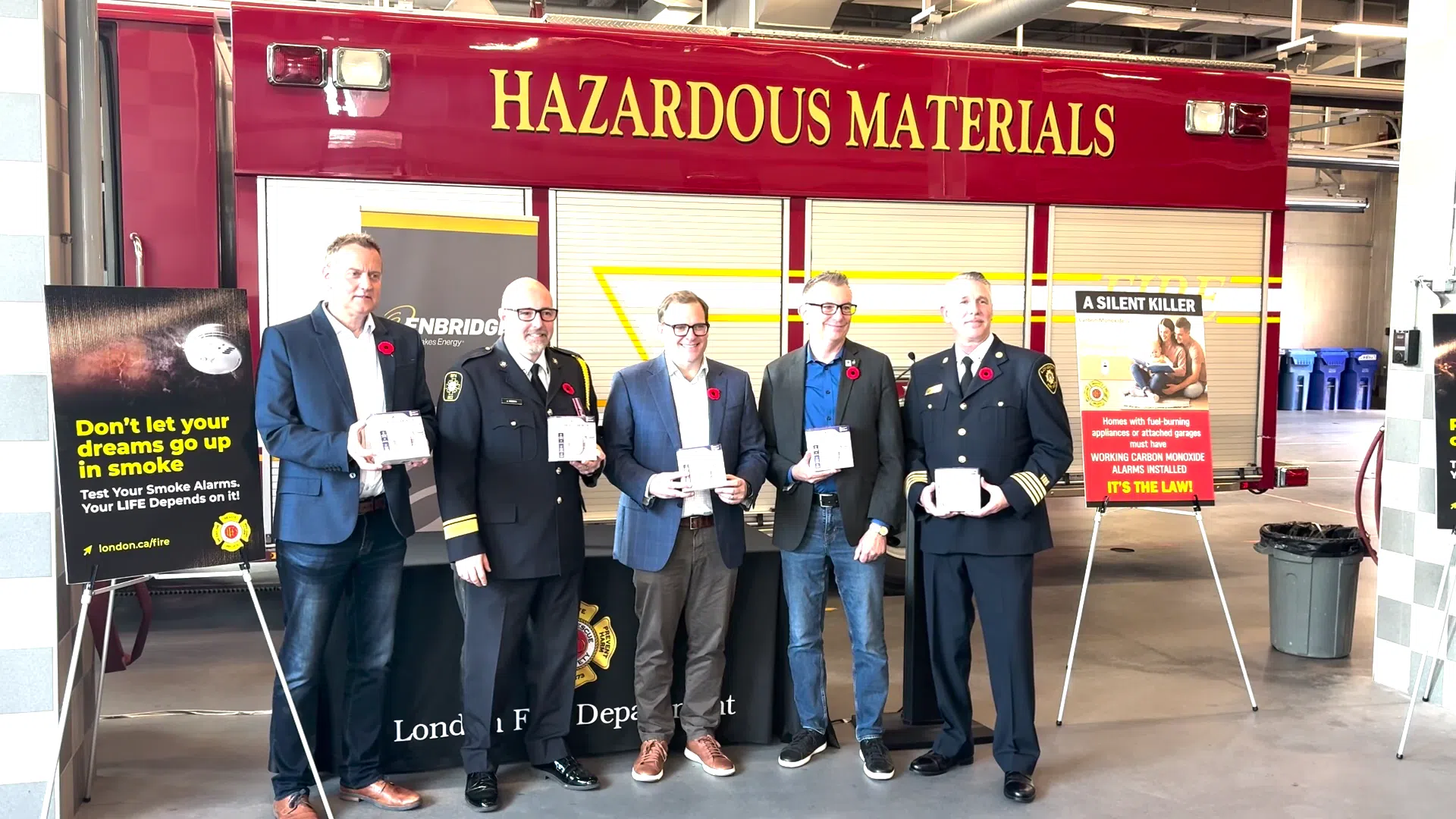 Five men—two in fire department dress uniforms and three in business attire—stand side by side in a fire station, each holding a boxed smoke or carbon monoxide alarm. They are positioned in front of a red Hazardous Materials fire truck. Safety posters on both sides of the group promote smoke alarm testing and carbon monoxide awareness. The setting reflects a public education event focused on home fire and CO safety.