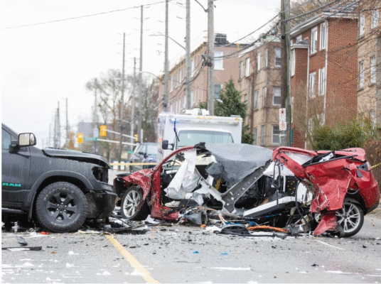 A severely damaged red car and a black pickup truck sit across a debris-covered Adelaide Street North following a major morning collision. Emergency vehicles and responders are visible behind the wreckage as the roadway remains closed for investigation and cleanup.