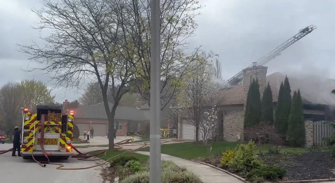 Firefighters operate an aerial ladder above a burning house while a fire engine in the foreground supplies water through connected hoses during a fire On Carnforth Road, London, ON.
